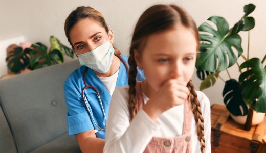 Family doctor at home in a medical protective mask listening to a child's lungs while sitting on a sofa in the living room. Patient examination, health care and medicine.